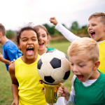 A group of children with cup prize standing outdoors on football pitch.